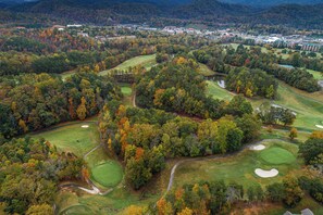 Eine Luftaufnahme eines Golfplatzes mit mehreren Grüns, Bahnen und Sandbunkern, umgeben von dichtem Wald und einer kleinen Stadt im Hintergrund.