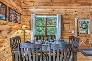 A wooden cabin interior with a dining table set for four, two chairs, a lamp, and a window with curtains. - Secluded Cabin with 2 En Suites & Hot Tub (Gatlinburg)