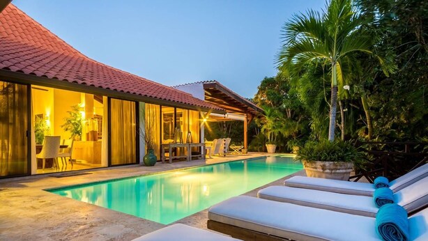Ein Pool mit Liegestühlen und einem überdachten Bereich, umgeben von viel Grün und einem Gebäude mit großen Fenstern.