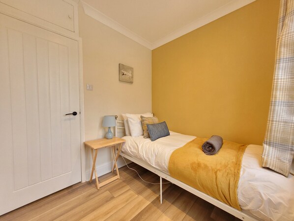 A single-bed room with a wooden floor, a white door, a bedside table with a lamp, and a yellow wall.