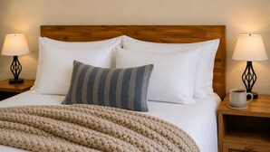 Ein ordentlich bezogenes Bett mit weißen und gestreiften Kissen, einer beigen Strickdecke sowie zwei Nachttische mit Lampen und einer Tasse.