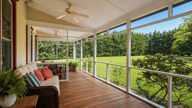 Eine überdachte Veranda mit Holzboden, einer Couch mit Kissen, einem Deckenventilator und Blick auf eine grüne Landschaft.