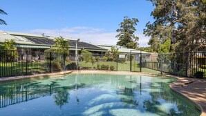 Una piscina con borde curvo, rodeada por una valla de metal negro, y contigua a un edificio con paneles solares.