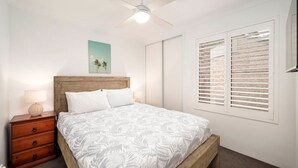 Un dormitorio con cama, cabecera de madera, mesitas de noche, ventilador de techo, ventana con persianas y un cuadro colgado en la pared.