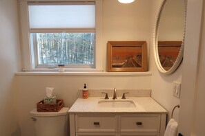 Casa de banho com sanita, lavatório e janela. Há um quadro emoldurado na parede acima do lavatório e um espelho na parede à direita.