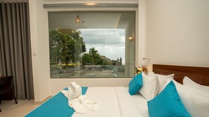 Habitación de hotel con un ventanal grande, cama con ropa de cama blanca y azul, cabecero de madera y un pequeño escritorio con una silla.