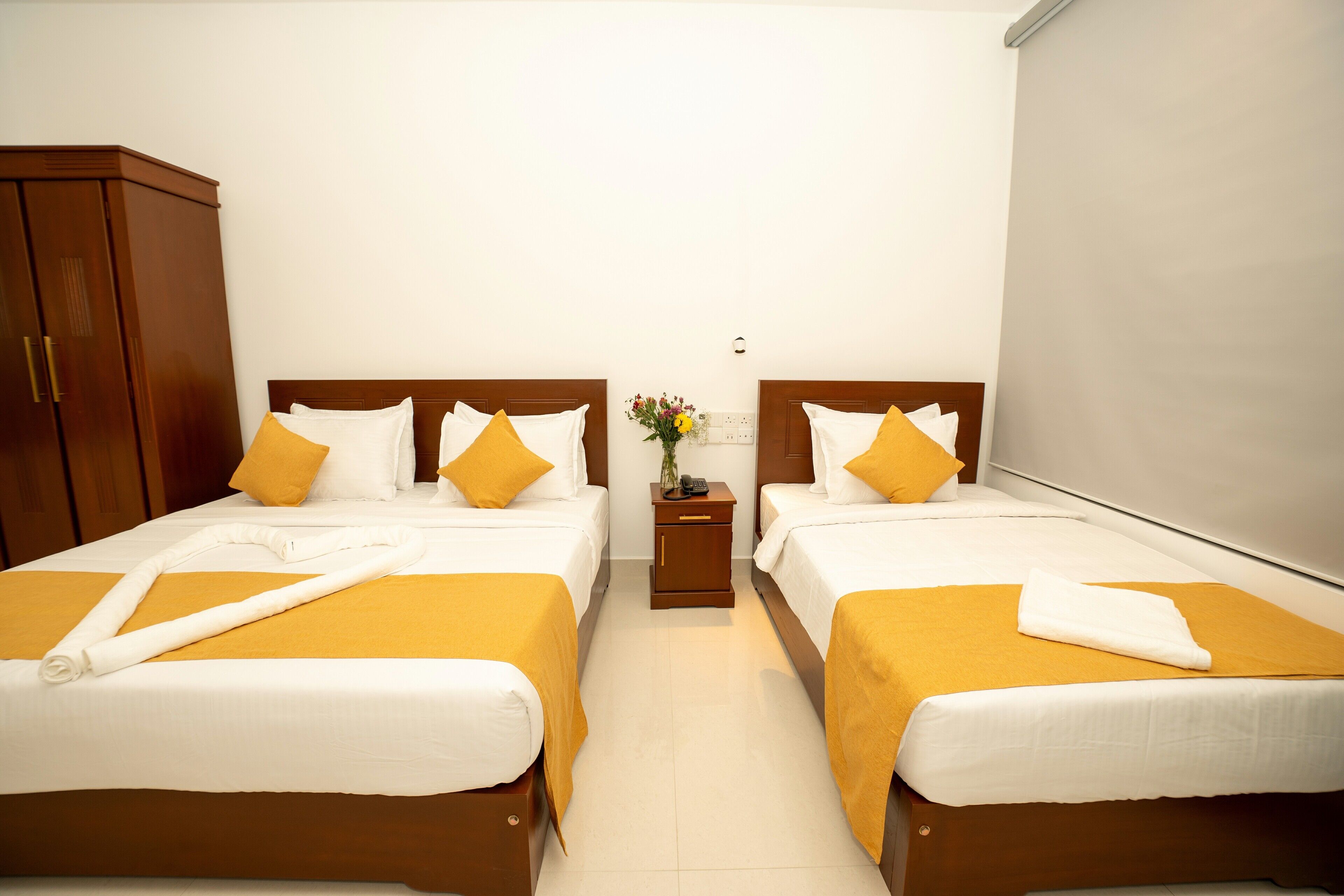 A hotel room with two beds, each with white linens and yellow accents, a wooden wardrobe, and a small bedside table with a vase of flowers.