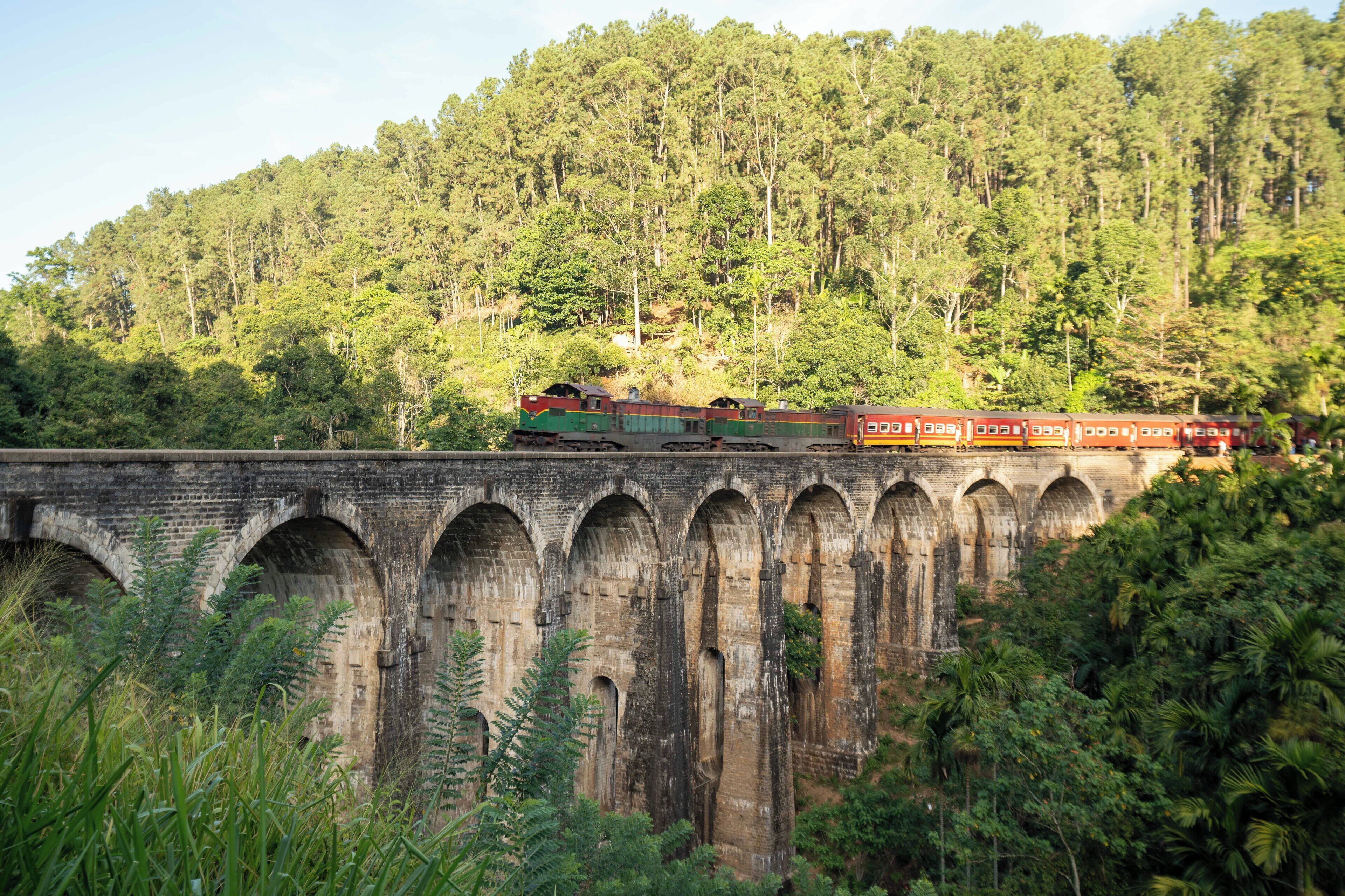 A train crossing a bridge with multiple arches, surrounded by dense forest.