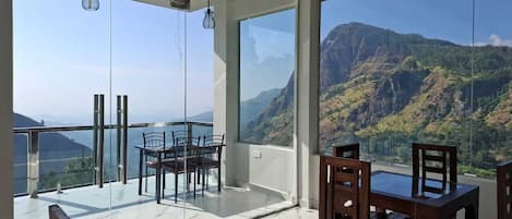 A balcony with a glass table and chairs, overlooking a mountainous landscape.