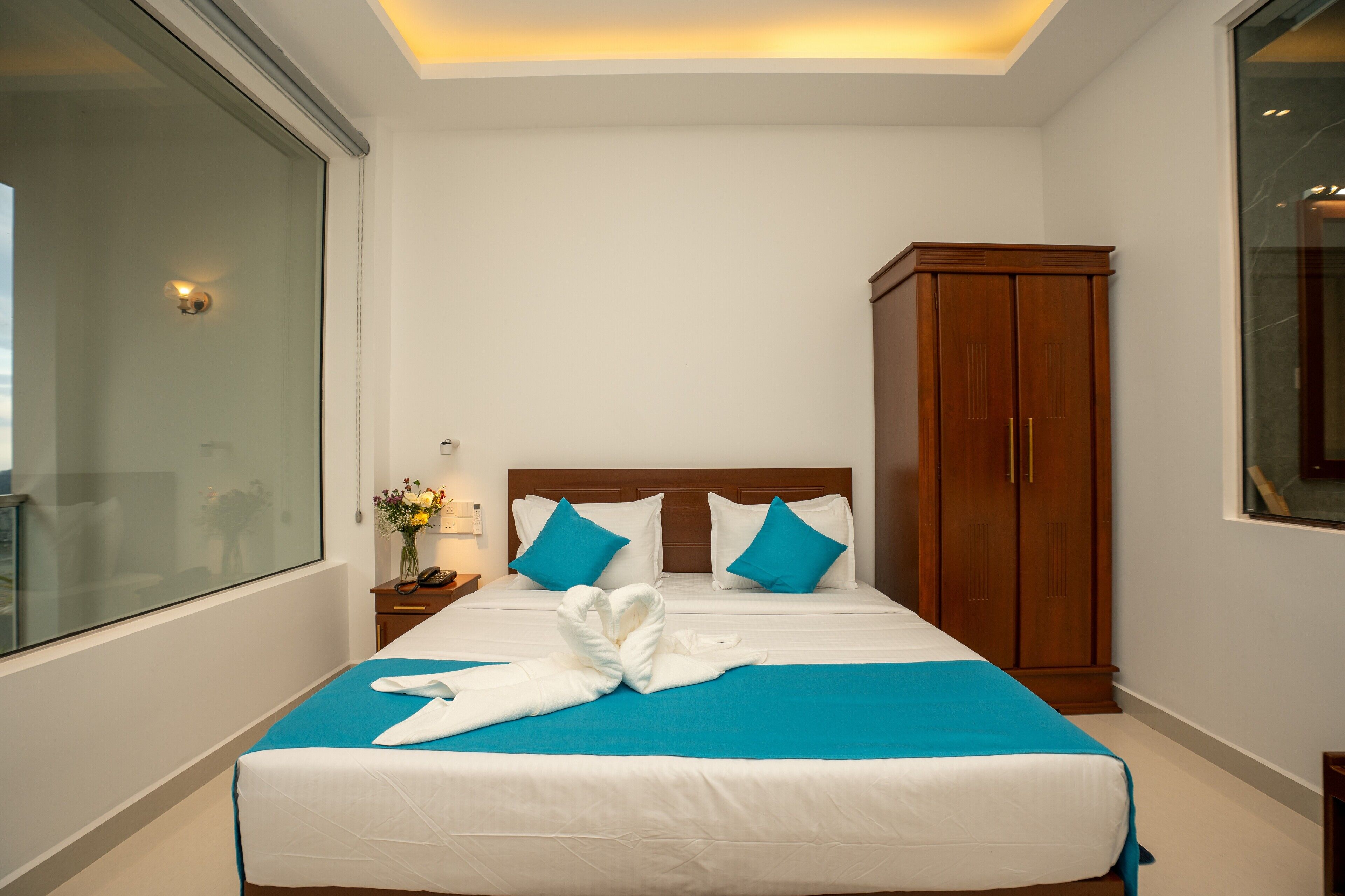 Ein ordentlich bezogenes Bett mit blauer Decke und weißen Laken, ein Holzschrank und ein Nachttisch mit Lampe und Blumen.