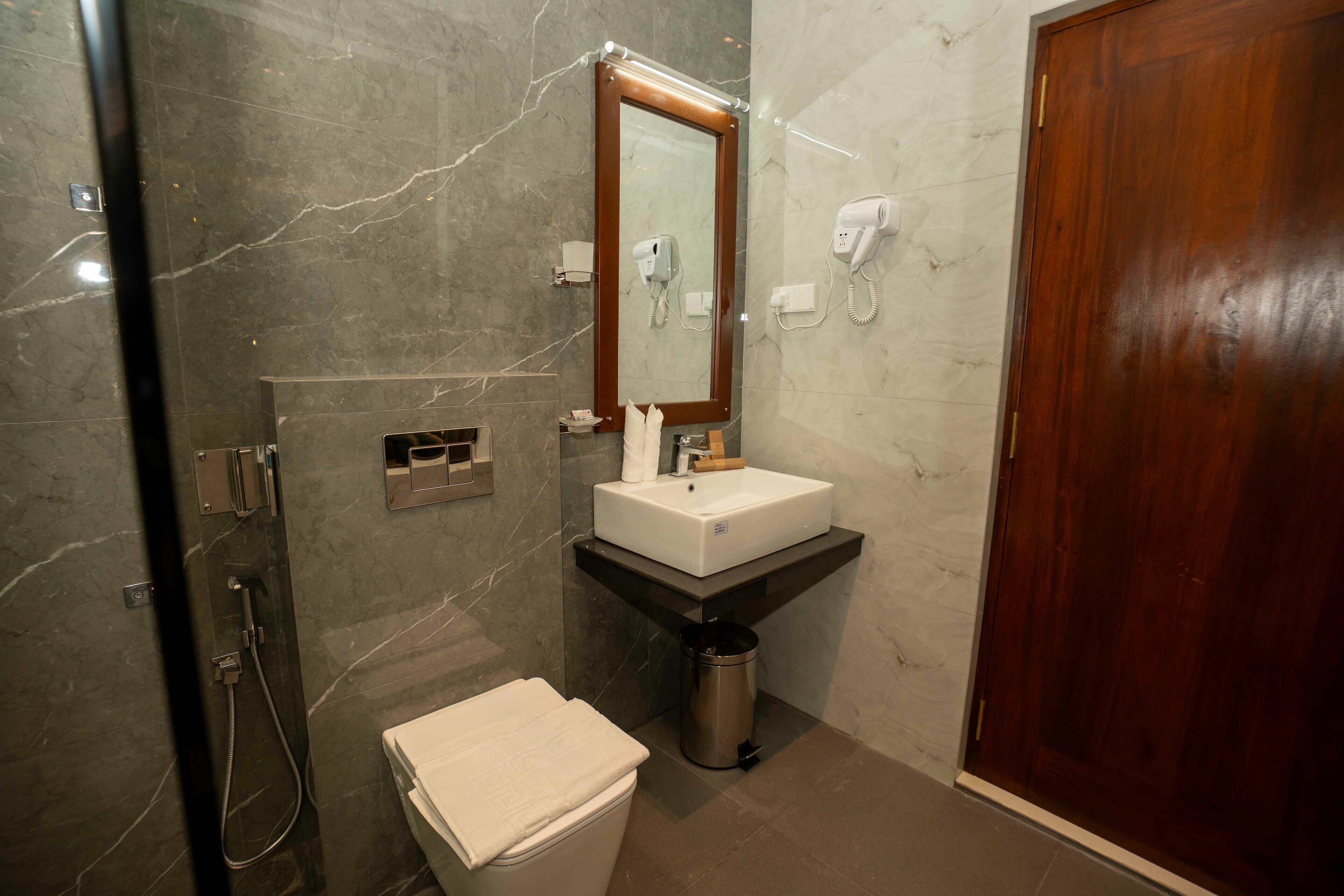 A bathroom with a marble wall, a wooden door, a sink with a mirror, a hairdryer, and a trash can.