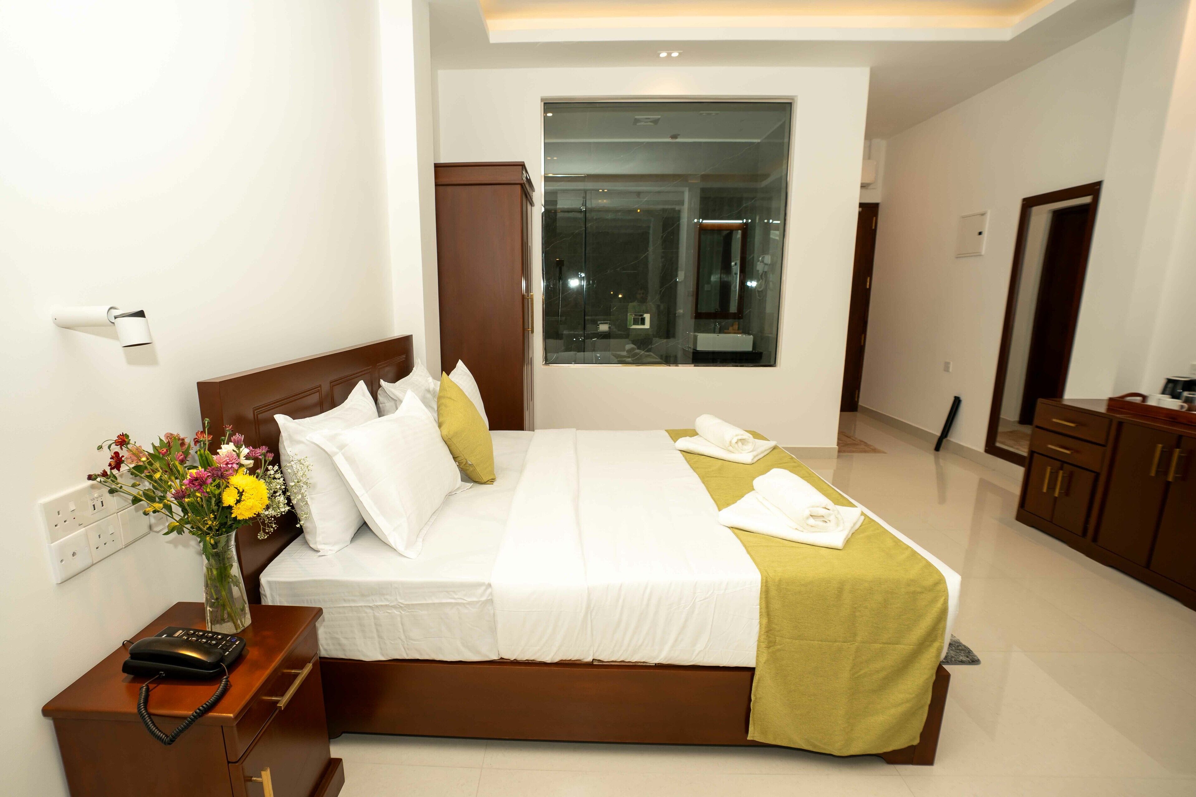 A hotel room with a large bed, bedside table with a phone, a vase of flowers, and a view of the city through a large window.