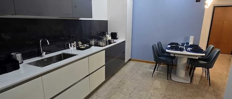 Une cuisine moderne avec des meubles blancs et noirs, un coin repas avec une table et des chaises, et un panier pour animaux de compagnie de couleur grise.