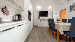 A modern kitchen with white cabinets, a coffee maker, and a kettle. There is a dining area with a table, chairs, and a TV mounted on the wall. - 3 bedroom lovely apartment in Maslenica (Maslenica)