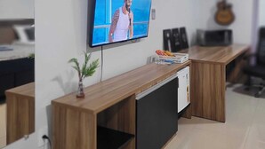 Un espacio de oficina moderno con un televisor de pantalla plana montado en la pared, un escritorio de madera, una pequeña planta en maceta y una guitarra al fondo.
