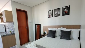 Habitación de hotel con cama, mesita de noche, espejo, pequeño escritorio con teléfono y vista a la cocina.