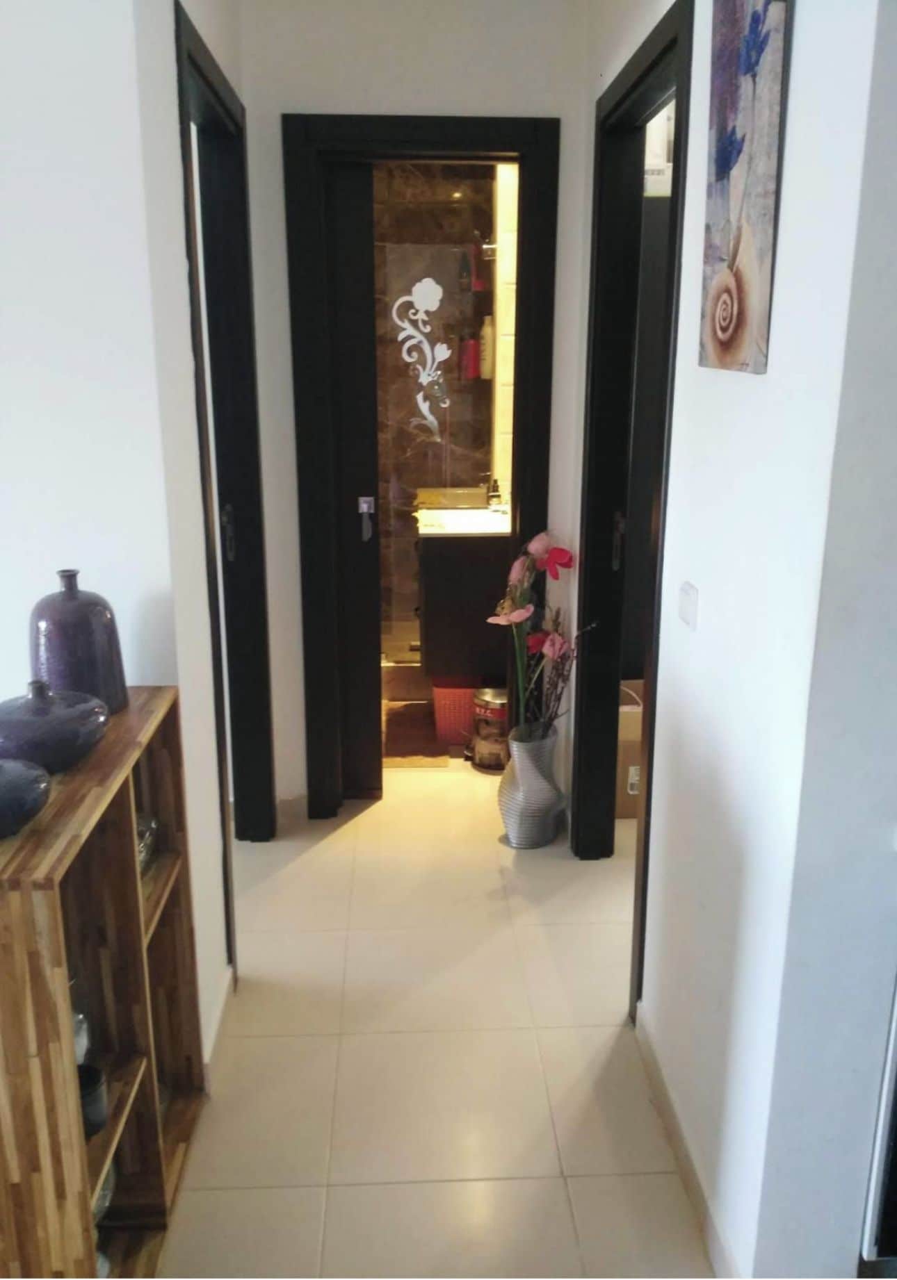 A narrow hallway with a wooden cabinet on the left and a door leading to a room with a bed and a vase of flowers.
