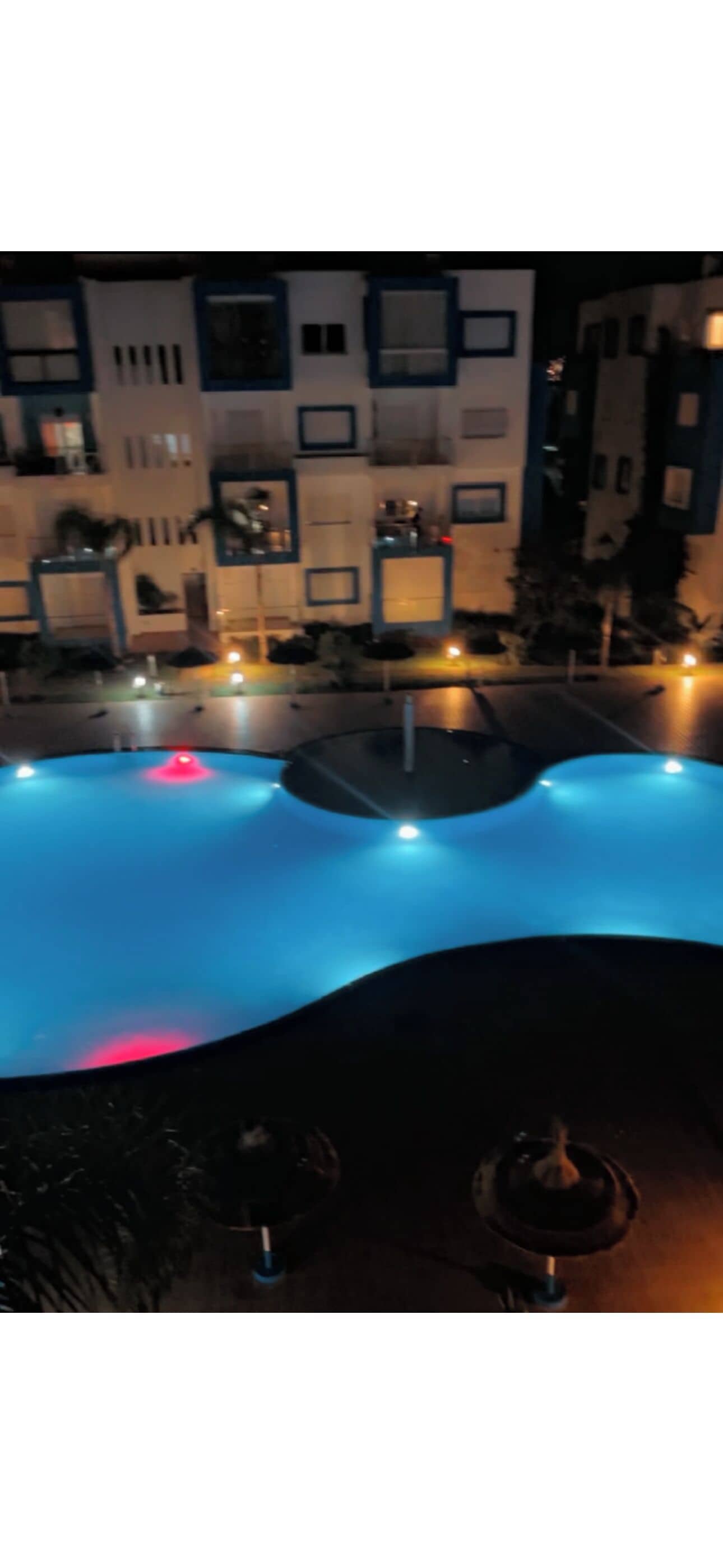 A swimming pool with a wavy edge and a fountain in the center, surrounded by buildings with balconies and outdoor lighting.