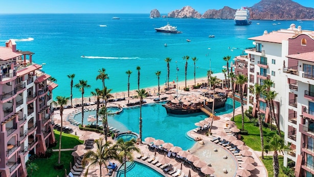 A resort with multiple swimming pools, palm trees, and a view of the ocean with boats.