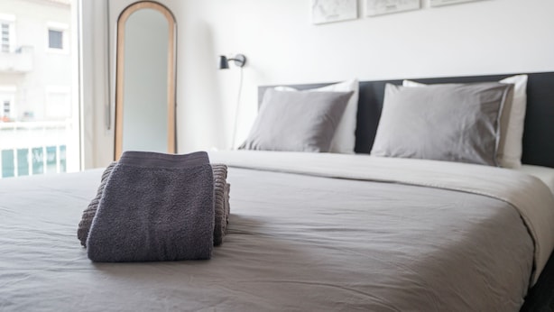A neatly made bed with a folded blanket, a bedside lamp, and a large mirror on the wall. - Central & Spacious 2BR in Almada • Easy access to Lisbon (Almada)