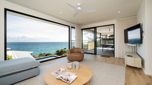 Ein modernes Wohnzimmer mit großem Fenster und Meerblick, Deckenventilator, Flachbildfernseher und Essbereich mit Tisch und Stühlen.