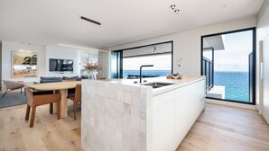 Eine moderne Küche mit einer großen Kochinsel, einem Essbereich mit Tisch und Stühlen, einem Flachbildfernseher und Meerblick durch große Fenster.