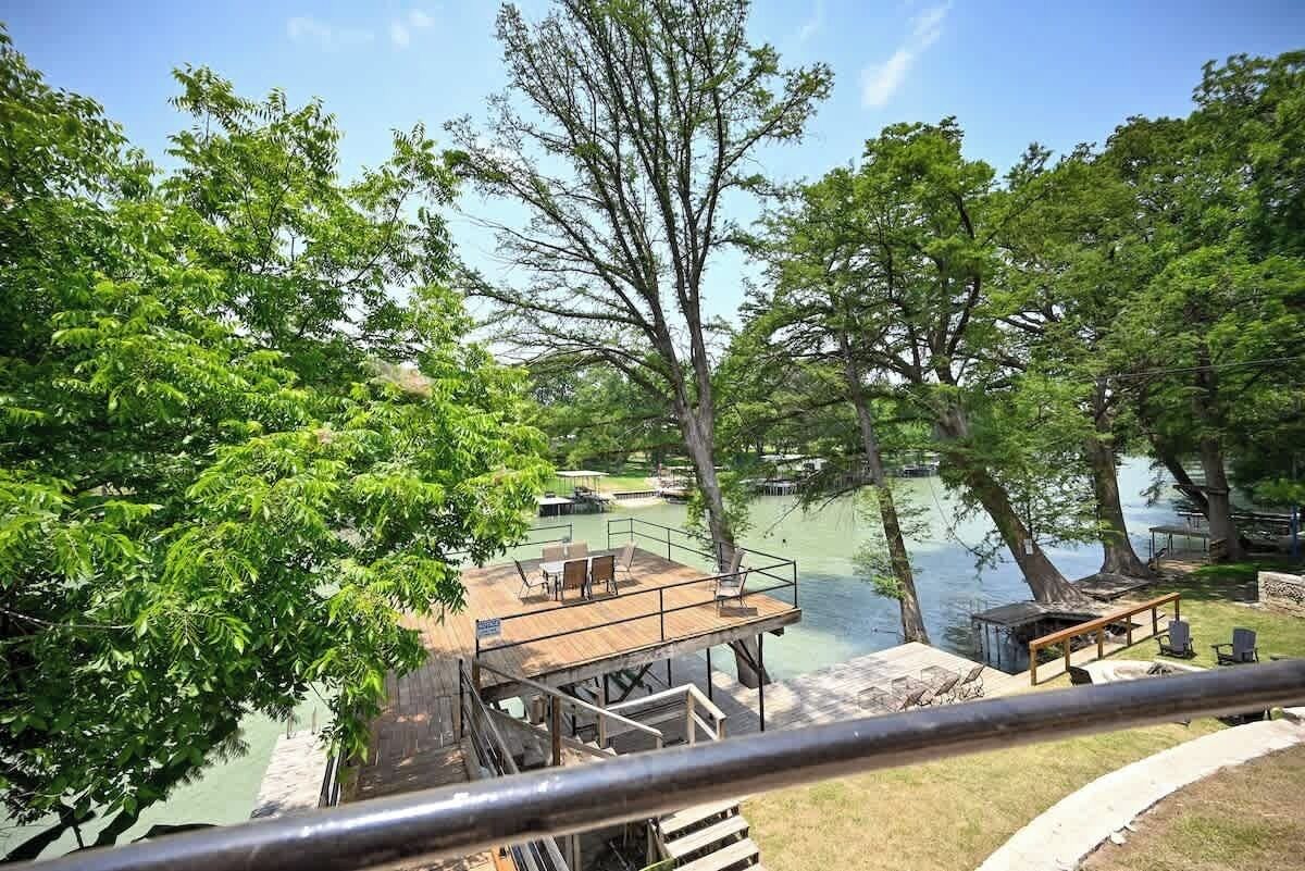 Una terrassa a la vora d'un llac amb seients exteriors i zona de barbacoa, envoltada d'arbres i amb vistes a una calma superfície d'aigua.