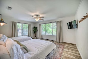 Un dormitorio con dos camas, ventilador de techo, televisor montado en la pared y ventanas amplias con cortinas.