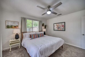 Un dormitorio con una cama, una mesita de noche, un ventilador de techo, una ventana con cortinas y dos cuadros enmarcados.
