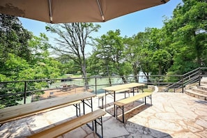 Mobiliario exterior con mesas y bancos, un patio de piedra y vistas a árboles y un cuerpo de agua.