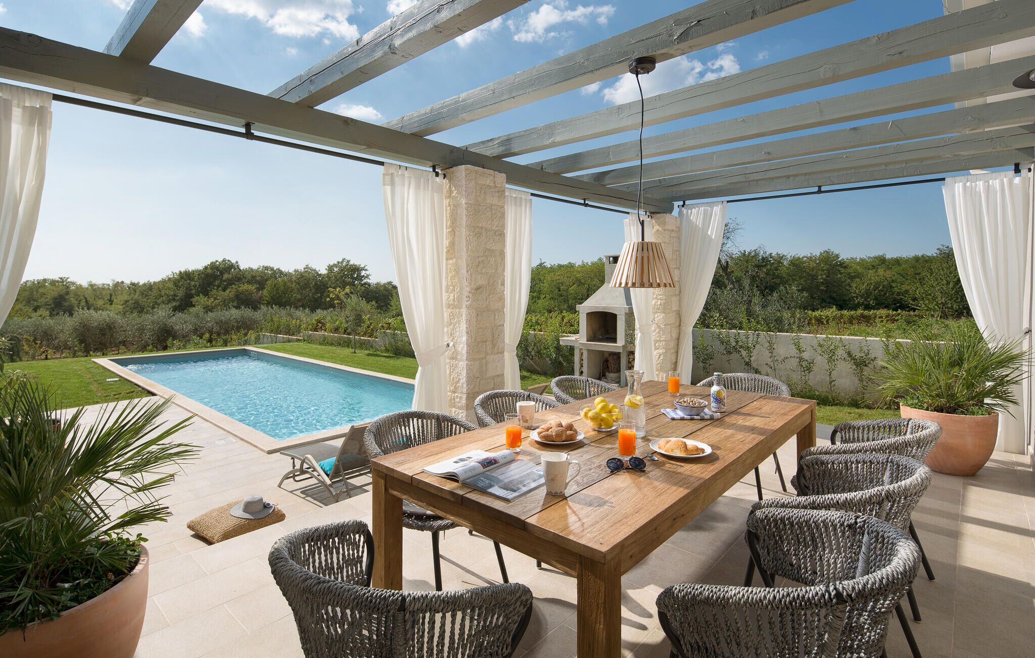 Ein Essbereich im Freien mit Blick auf einen Pool, Korbstühle, ein Holztisch und ein Pizzaofen.
