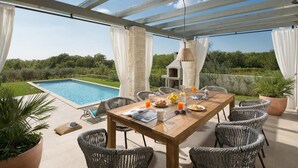 An outdoor dining area with a pool view, wicker chairs, a wooden table, and a pizza oven.