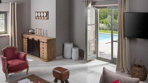 A spacious living room with a wooden cabinet, a red armchair, a leather ottoman, and a large window with a view of a swimming pool.