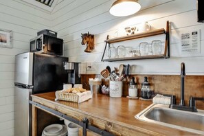 A kitchen with a wooden countertop, a stainless steel refrigerator, a microwave, and a sink. There are utensils in a container, a jar of utensils, and a shelf with glassware.