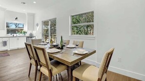 Un coin repas avec une table en bois, dressée pour quatre personnes, entourée de chaises. Des fenêtres offrent une vue sur la verdure environnante.