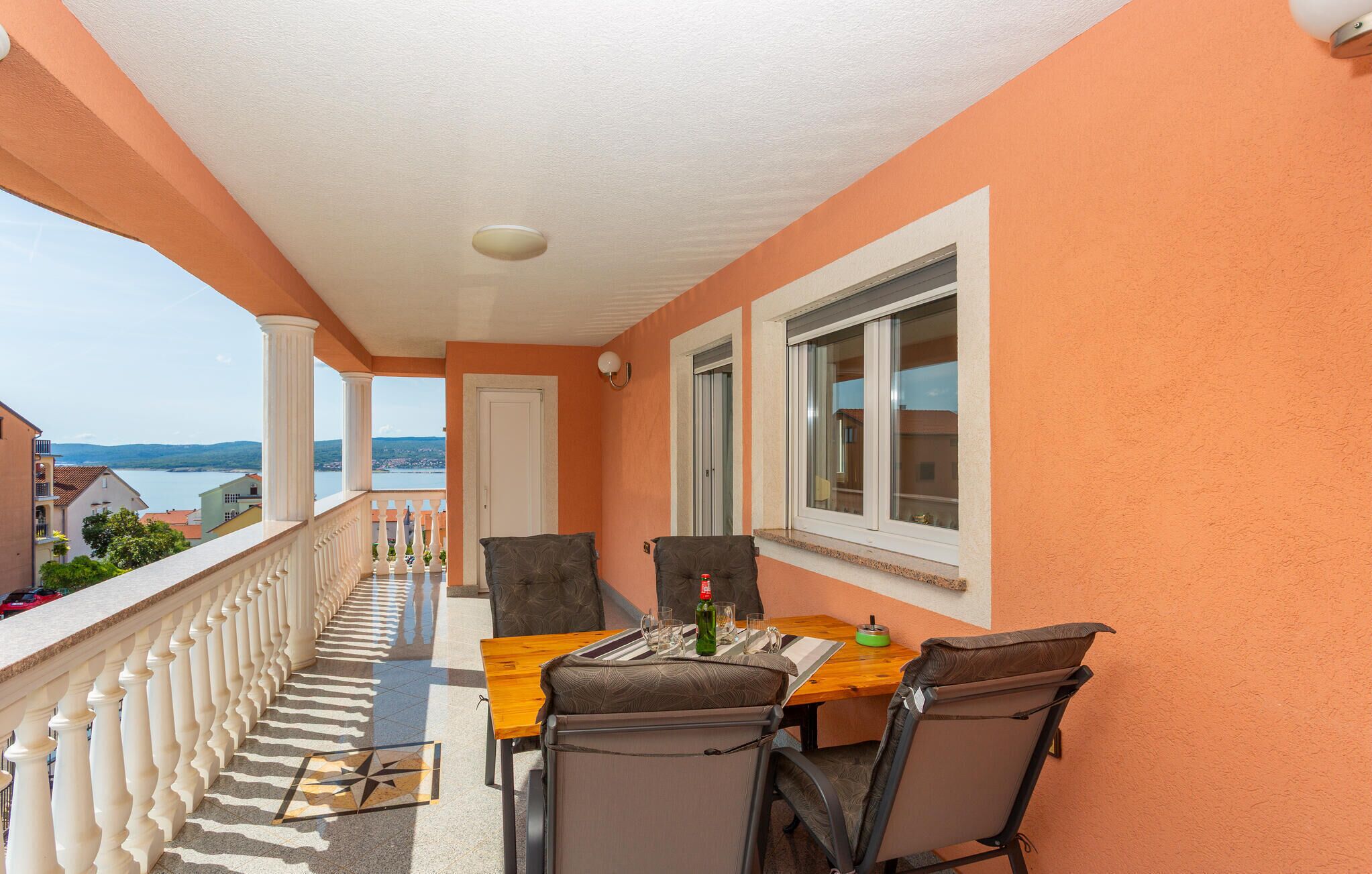 A balcony with a table and chairs, a view of the sea, and a building with orange walls.