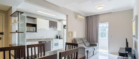 Una cocina moderna con armarios blancos, microondas empotrado y cocina a gas. Hay una zona de comedor con mesa de vidrio y sillas de madera. Un sofá gris se encuentra cerca de un amplio ventanal con cortinas.