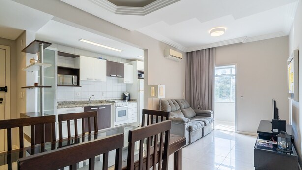 A modern kitchen with white cabinets, a built-in microwave, and a gas stove. There is a dining area with a glass table and wooden chairs. A grey sofa is positioned near a large window with curtains. - Apto 2 Qtos c/ Churrasq. a 650m da praia ESP0301 (Florianópolis)