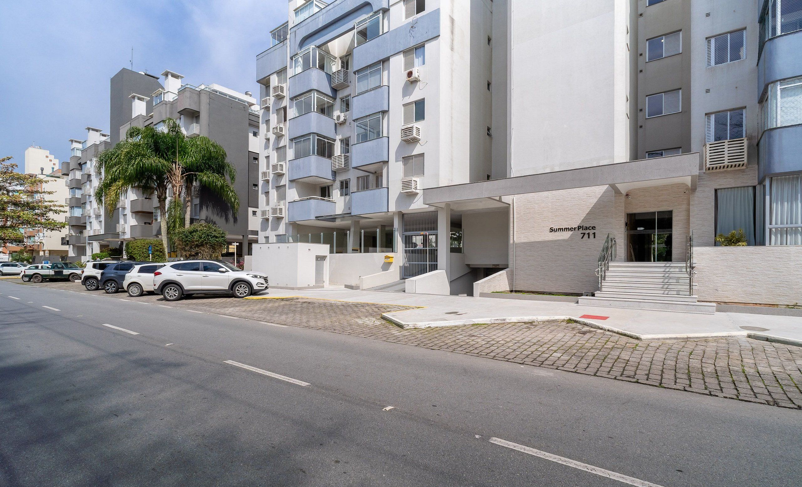 Un moderno edificio de apartamentos con fachada plana, varios balcones y un estacionamiento con varios automóviles.