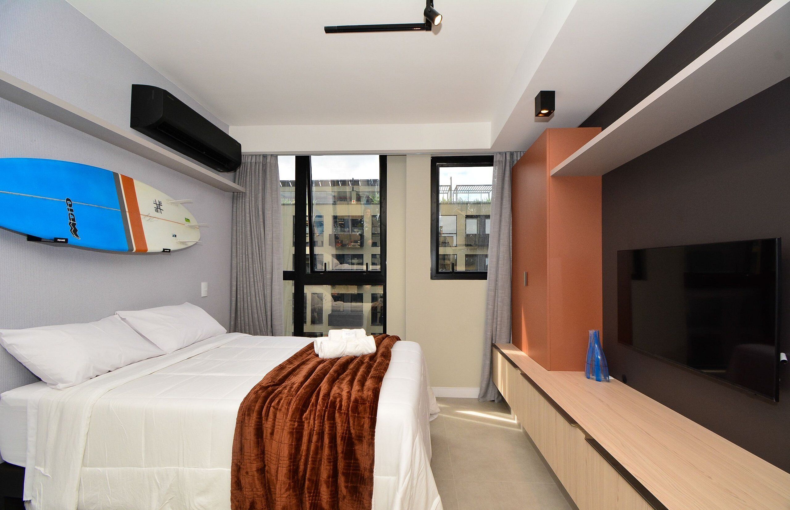 A hotel room with a bed, a TV, a surfboard, and a window with a city view.