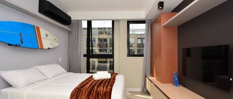 A hotel room with a bed, a TV, a surfboard, and a window with a city view.