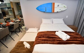Hotelkamer met een bed, een bruine deken, een wit kussen, een blauw-witte surfplank aan de muur, en een eethoek met een tafel gedekt voor twee personen.