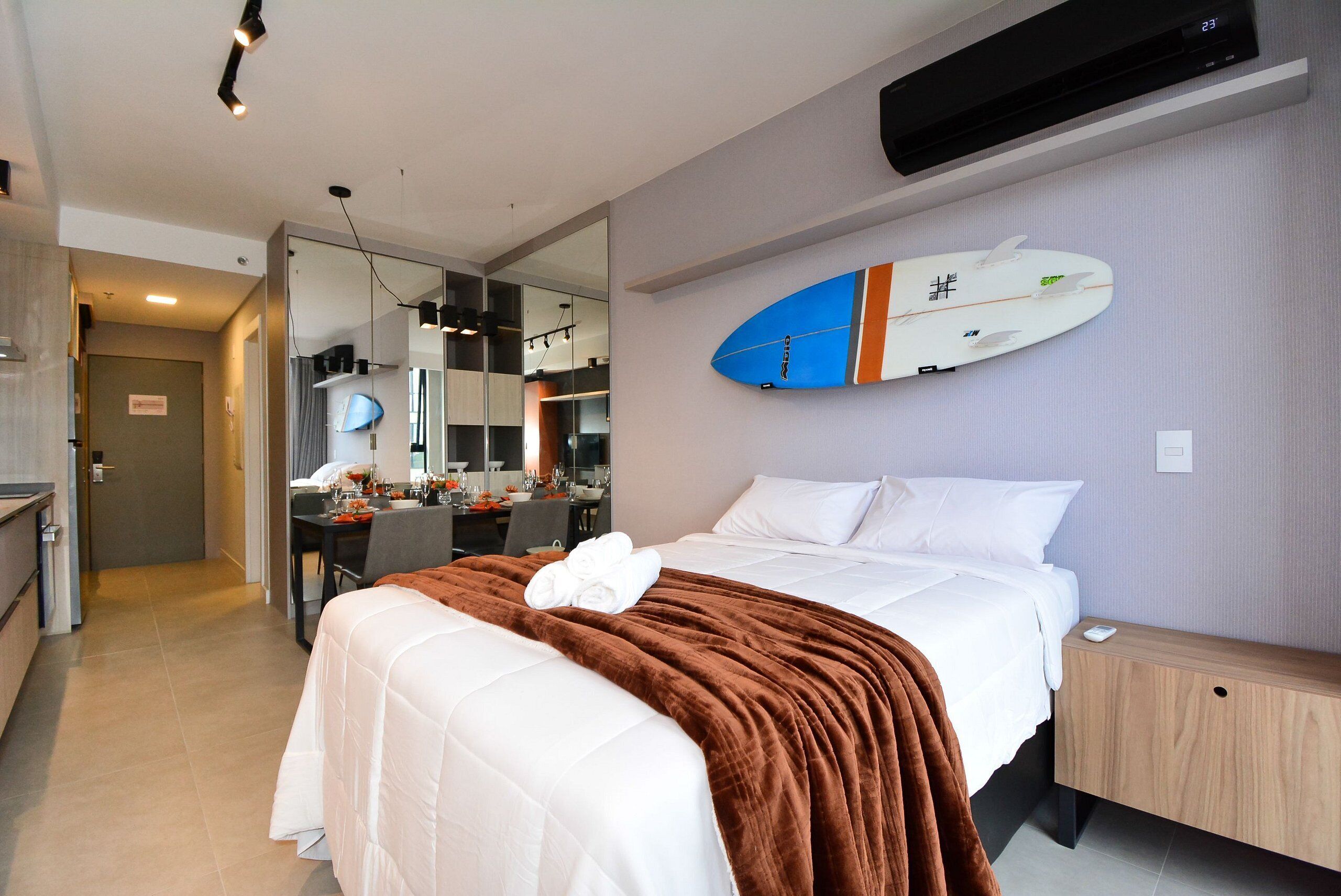 Ein Hotelzimmer mit Bett, Nachttisch aus Holz, Spiegel, Surfbrett an der Wand und einer Küchenzeile.