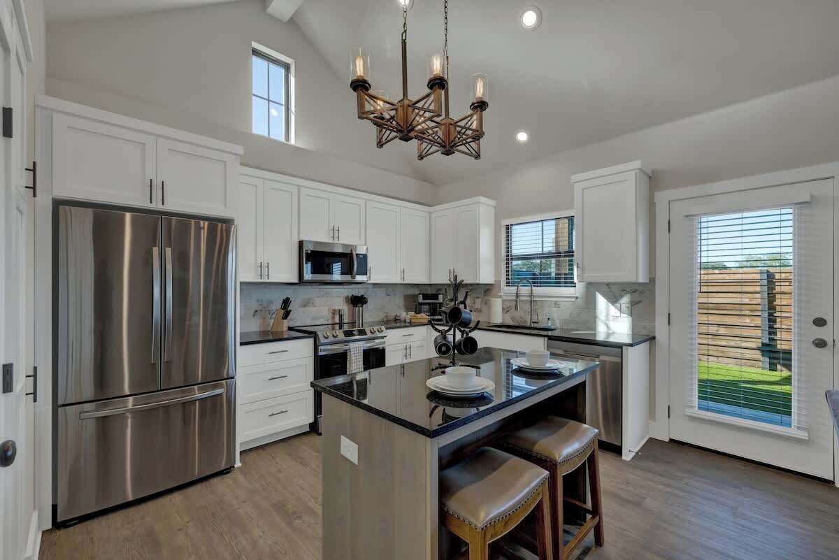 A modern kitchen with white cabinets, stainless steel appliances, a central island with stools, and a chandelier.