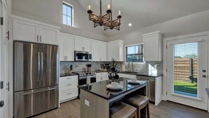 A modern kitchen with white cabinets, stainless steel appliances, a central island with stools, and a chandelier.