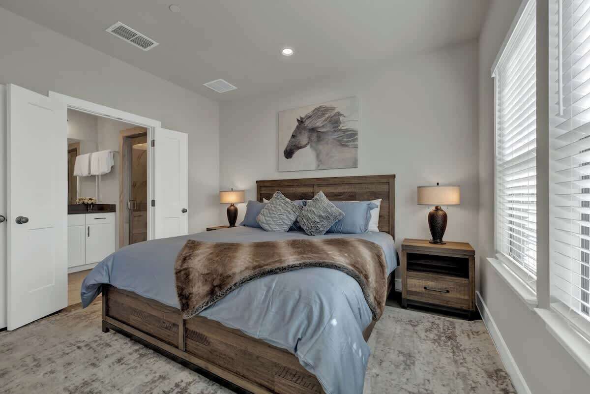 A bedroom with a wooden bed, two bedside tables with lamps, a large window with blinds, a door leading to a bathroom, and a horse-themed artwork on the wall.