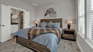 A bedroom with a wooden bed, two bedside tables with lamps, a large window with blinds, a door leading to a bathroom, and a horse-themed artwork on the wall.