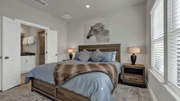 A bedroom with a wooden bed, two bedside tables with lamps, a large window with blinds, a door leading to a bathroom, and a horse-themed artwork on the wall.