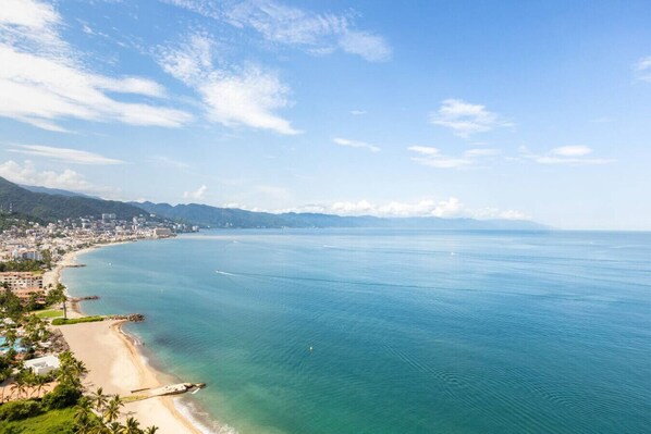 A coastal cityscape with a beach, clear blue sea, and distant mountains.