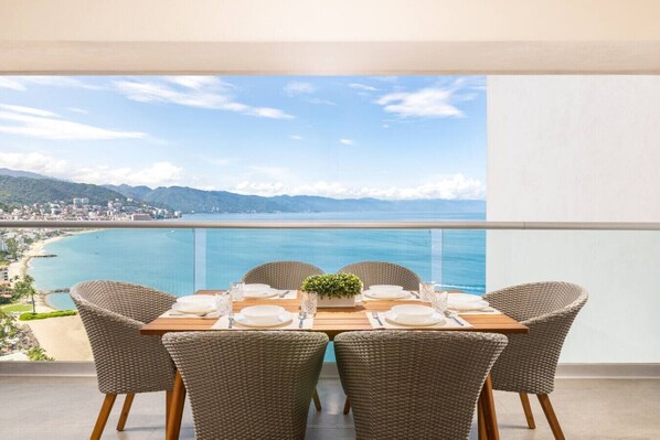 A dining table set for four with wicker chairs, overlooking a coastal view.